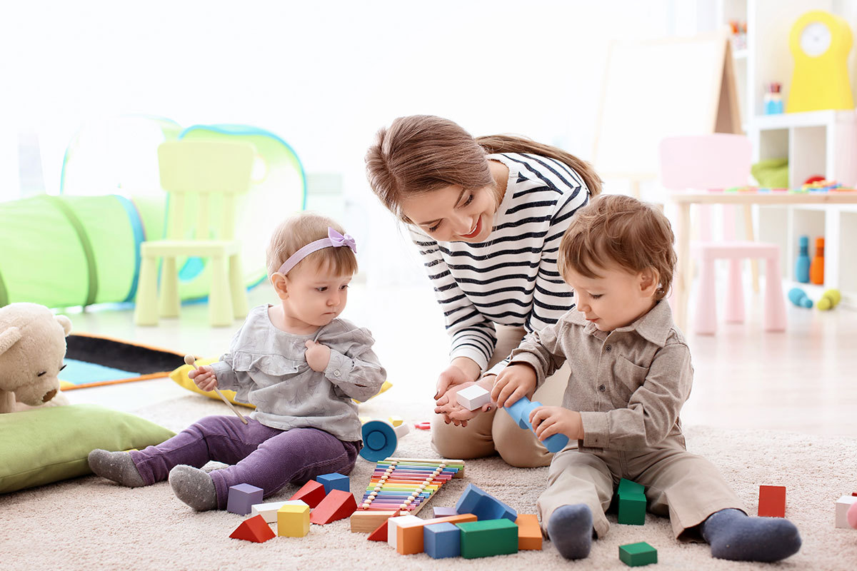 young smiling white nanny sits on floor with baby girl and a happy toddler boy playing with building blocks